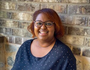 A photo of black fantasy author D. Hale Rambo. She is sitting next to a brown brick wall smiling at the camera. She is wearing a blue top with white spots and glasses.